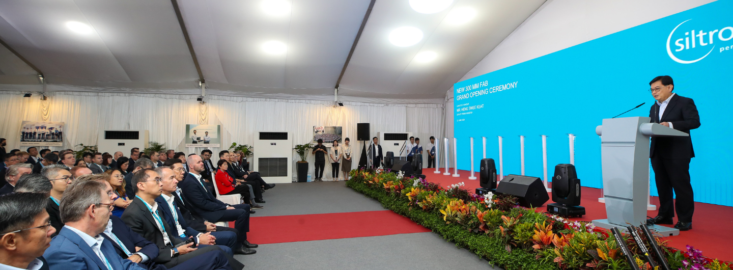 Audience listens as speaker at podium with "Siltronic" logo, "Grand Opening Ceremony" on a blue screen.